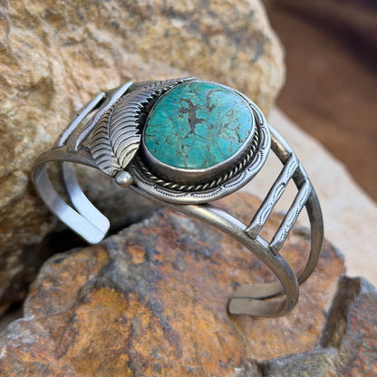 sterling silver cuff with large turquoise cabochon set in bezel surrounded by rope twist and other ornate details. it is sitting on a rock.