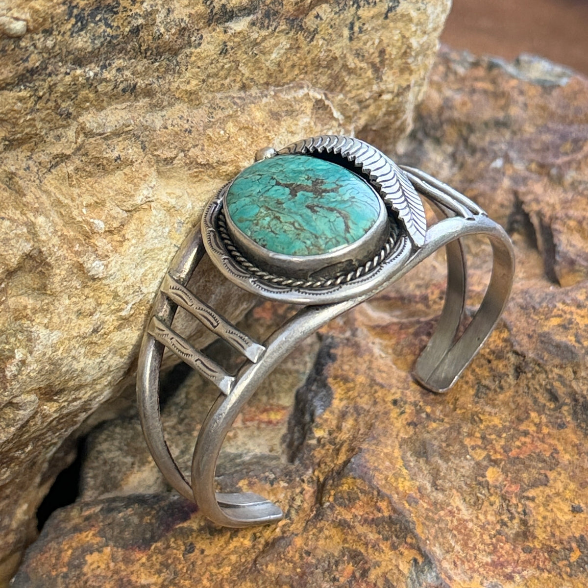 sterling silver cuff with large turquoise cabochon set in bezel surrounded by rope twist and other ornate details. it is sitting on a rock.