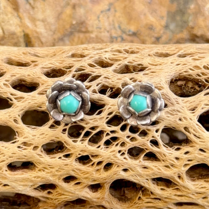A pair of silver-toned flower-shaped stud earrings with round turquoise centers, displayed on a textured beige bone or driftwood surface.