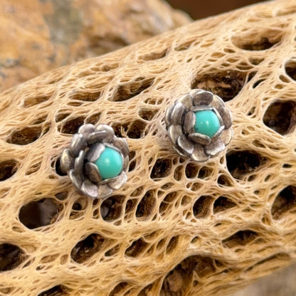 A pair of silver-toned flower-shaped stud earrings with round turquoise centers, displayed on a textured beige driftwood surface.