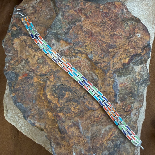 A colorful geometric link bracelet with silver-toned metal and multicolored inlay tiles, displayed diagonally on a textured brown and orange rock.