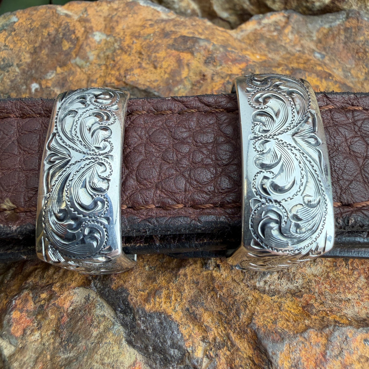 Silver belt buckle with intricate designs on a brown leather belt against a rocky background