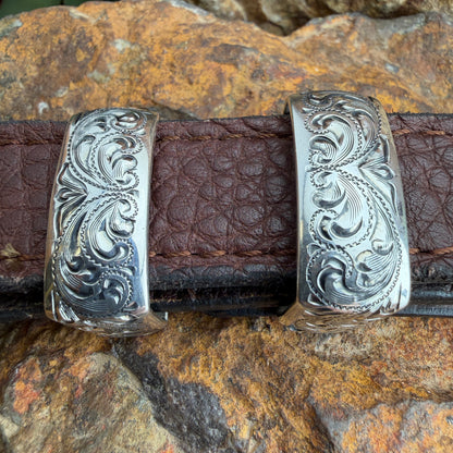 Silver belt buckle with intricate designs on a brown leather belt against a rocky background