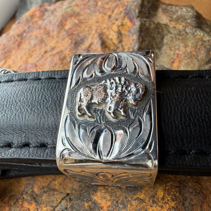 Silver belt buckle with engraved design on a black belt against a stone background