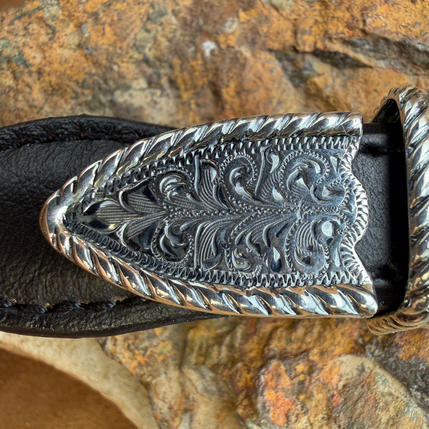 Silver belt buckle with intricate designs on a black leather strap against a stone background