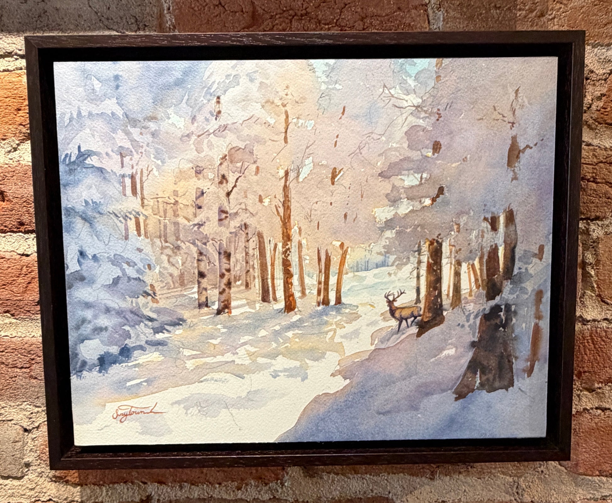 Framed painting of a winter landscape with a deer on a brick wall.
