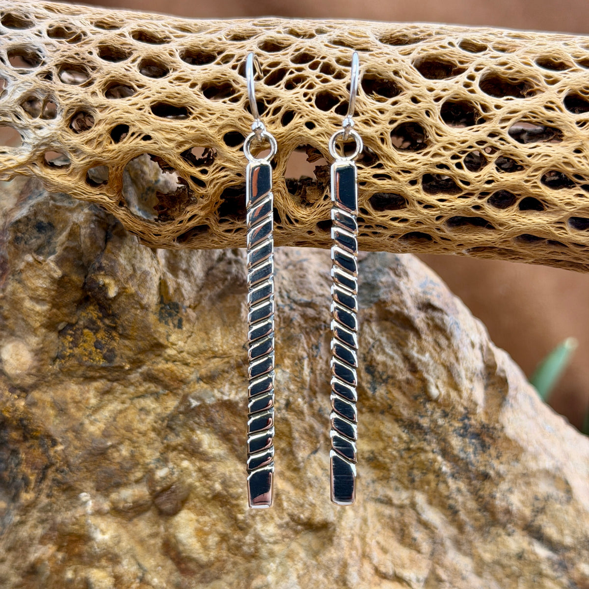 elongated rectangle earrings with diagonal geometric line pattern by artie yellowhorse hanging from a piece of driftwood